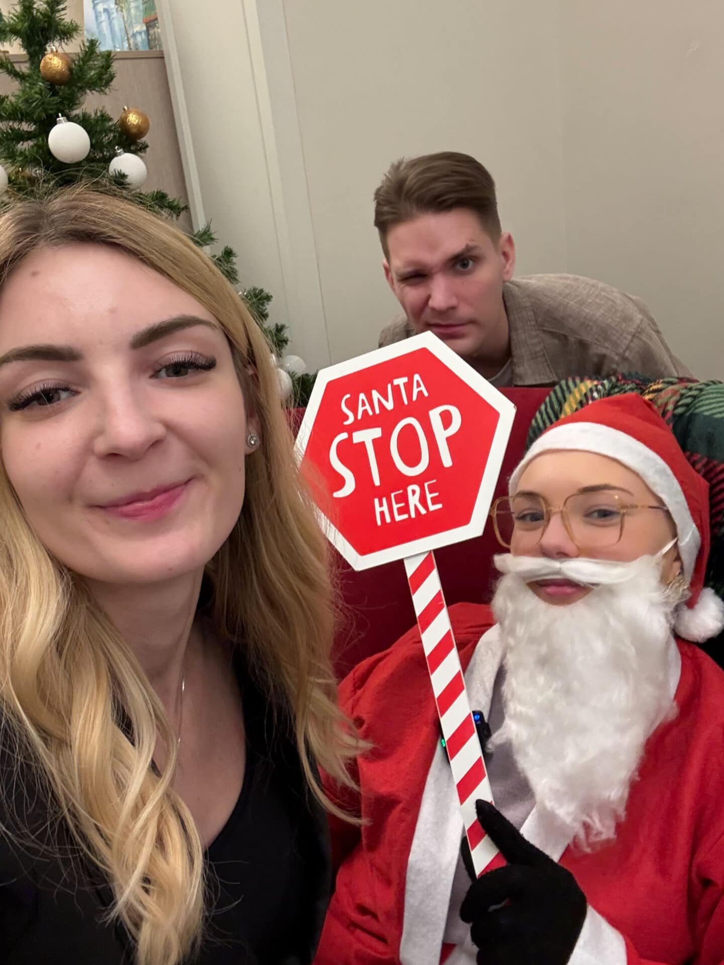 1. Draudzīga Ziemassvētku ballītes bilde ar cilvēkiem un Santa Claus ar "Santa Stop Here" bāru, svētku dekorācijām un Ziemassvētku eglīti.