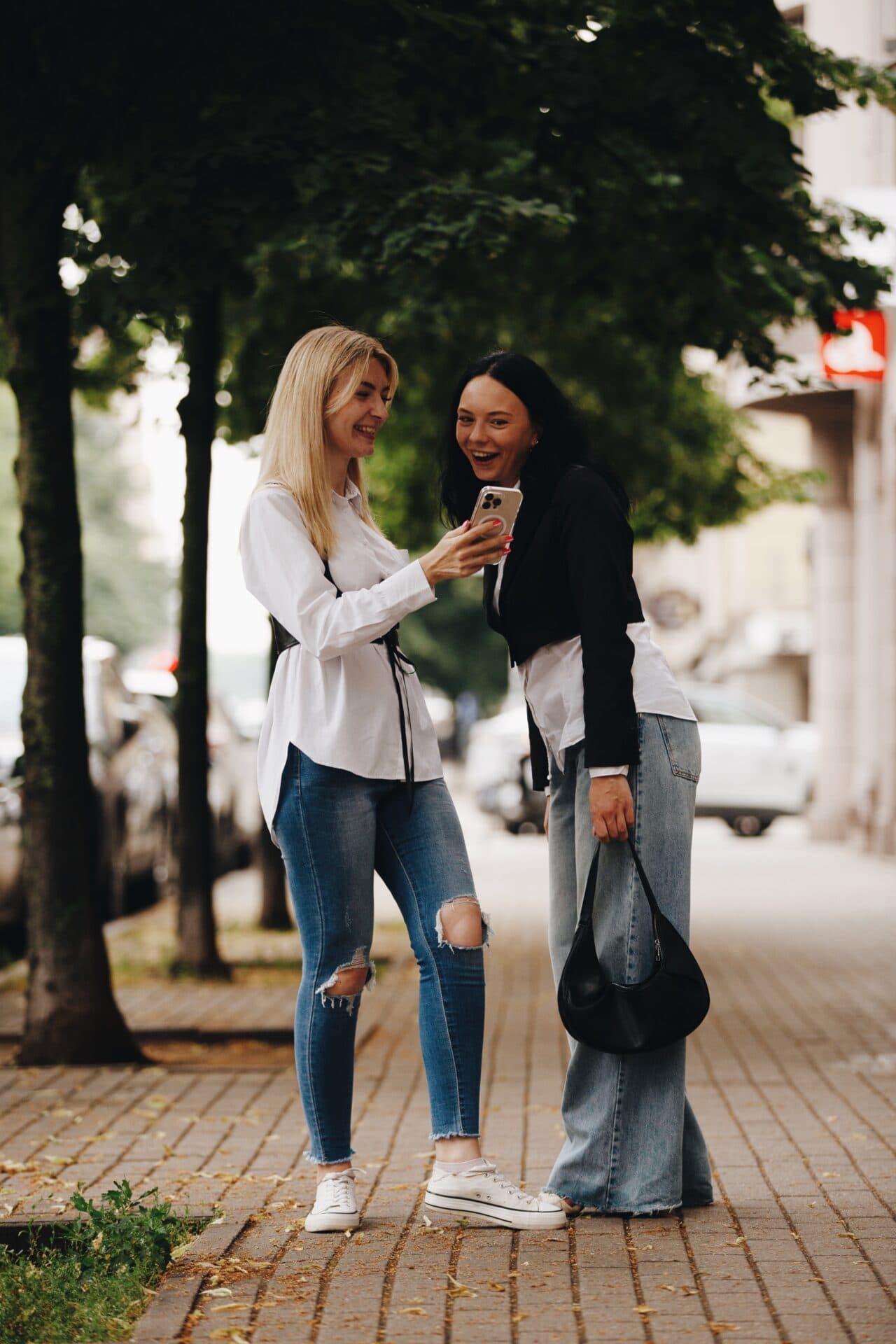 Svešinieču apbrīnojama draudzība un dzīvesprieks, pētot telefonu kopā pilsētas ielā, ar zaļu apstādījumu un modernu apkārtni fonā, veidojot draudzīgas un modernās dzīves ainu.