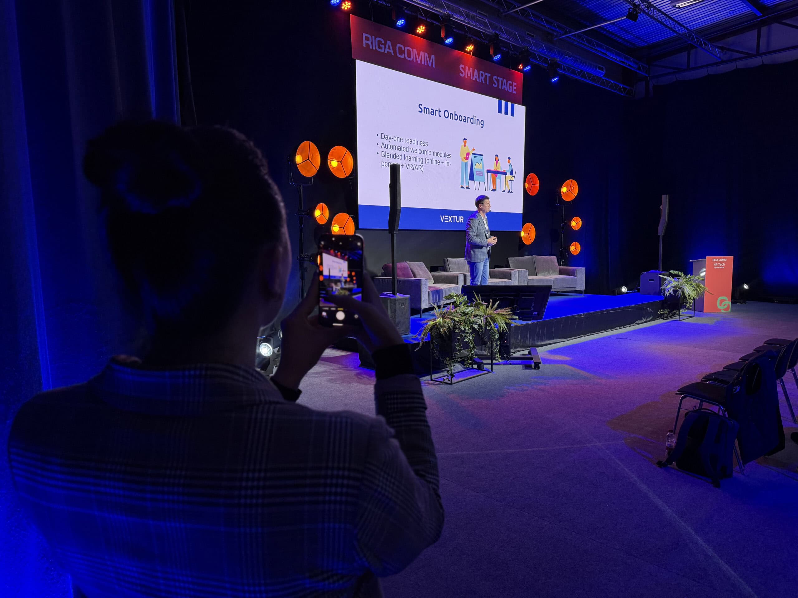 Person fotografē prezentāciju par "Gudru iecelšanu" konferencē Rīgā, ar skatu uz skatuvi un prezentācijas ekrānu.